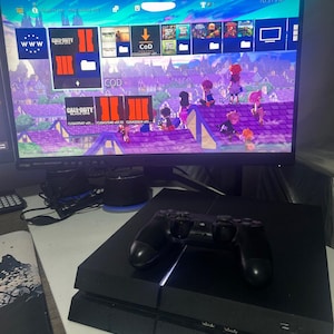May include: A black PlayStation 4 gaming console with a matching black wireless controller resting on top. The console is positioned in front of a curved computer monitor displaying a video game console interface with various game icons and titles.