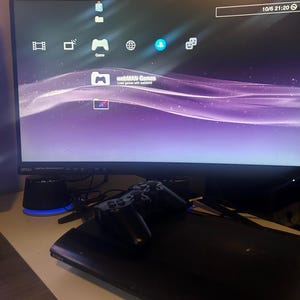 Könnte beinhalten: Eine schwarze Sony PlayStation 3 Konsole mit einem passenden schwarzen kabellosen Controller, der darauf liegt. Die Konsole ist mit einem gebogenen MSI-Monitor verbunden, der die PlayStation 3 XrossMediaBar-Oberfläche mit violettem Wellenhintergrund zeigt.
