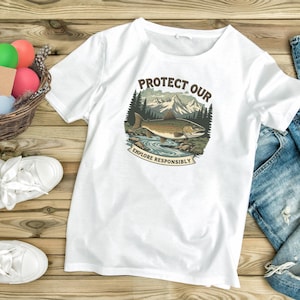 Puede incluir: Camiseta blanca con un diseño gráfico circular que presenta un pez, montañas y árboles. El texto dice "PROTECT OUR" encima de la imagen y "EXPLORE RESPONSIBLY" debajo. La camiseta está hecha de un material suave.