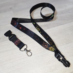 Op de afbeelding: Zwart koord met een herhalend patroon van hondengezichten en sterren in goud, rood en groen. Het bevat een zwarte plastic sluiting en een bandje met de tekst "SPACE WOLF".