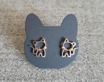 May include: Rose gold cat-shaped stud earrings on a black cat-shaped earring card. The earrings are a simple outline of a cat with a tail and are made of a shiny metal. The background is a grey, textured surface.