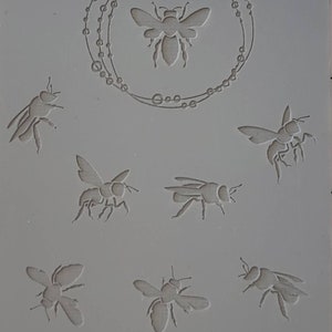 Puede incluir: Un molde de silicona con siete diseños de abejas. Las abejas están en varias posiciones, incluyendo volando, arrastrándose y descansando. El molde también presenta un círculo de pequeños puntos alrededor de una sola abeja en el centro. El texto "Cabtopia.com 100133" está impreso en la parte inferior del molde.