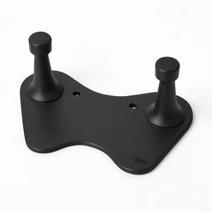 Puede incluir: Un soporte negro mate con dos postes verticales y una base curva. Los postes son cilíndricos con una parte superior redondeada. La base tiene una ligera curva y dos pequeños agujeros. El soporte está diseñado para sostener un objeto.
