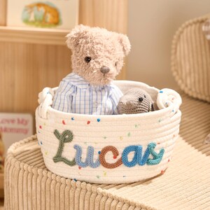 May include: A cream-colored rope basket with the name "Lucas" embroidered in colorful letters. The basket contains a teddy bear wearing a blue and white striped shirt and a small gray stuffed animal. The basket is on a corduroy surface.