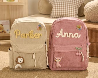 May include: Two corduroy backpacks, one beige and one pink, are displayed. The beige backpack has the name "Parker" in yellow, a lion applique, and a flower. The pink backpack has the name "Anna" in white, a bear applique, and a flower.