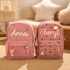 May include: Two pink corduroy backpacks. One backpack has the name "Anna" in white script, a flower, a bear, and cherries. The other backpack has the name "Sheryl" in white script, a flower, and strawberry designs.