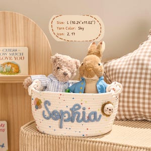 May include: A cream-colored rope basket with the name "Sophia" in blue yarn, holding a teddy bear and a rabbit plush toy. The basket is approximately 10.24 inches long and 11.02 inches wide. A decorative flower and lion icon are attached to the basket.