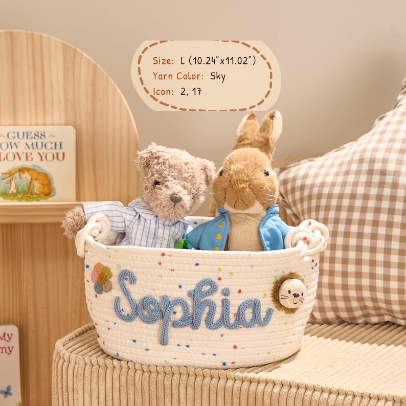 May include: A cream-colored rope basket with the name "Sophia" in blue yarn, holding a teddy bear and a rabbit plush toy. The basket is approximately 10.24 inches long and 11.02 inches wide. A decorative flower and lion icon are attached to the basket.