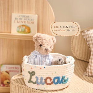 May include: A small, cream-colored rope basket with the name "Lucas" embroidered in green and blue yarn. The basket contains a teddy bear wearing a blue and white checkered shirt and a small gray crocheted animal. The basket measures 4.72" x 8.66".