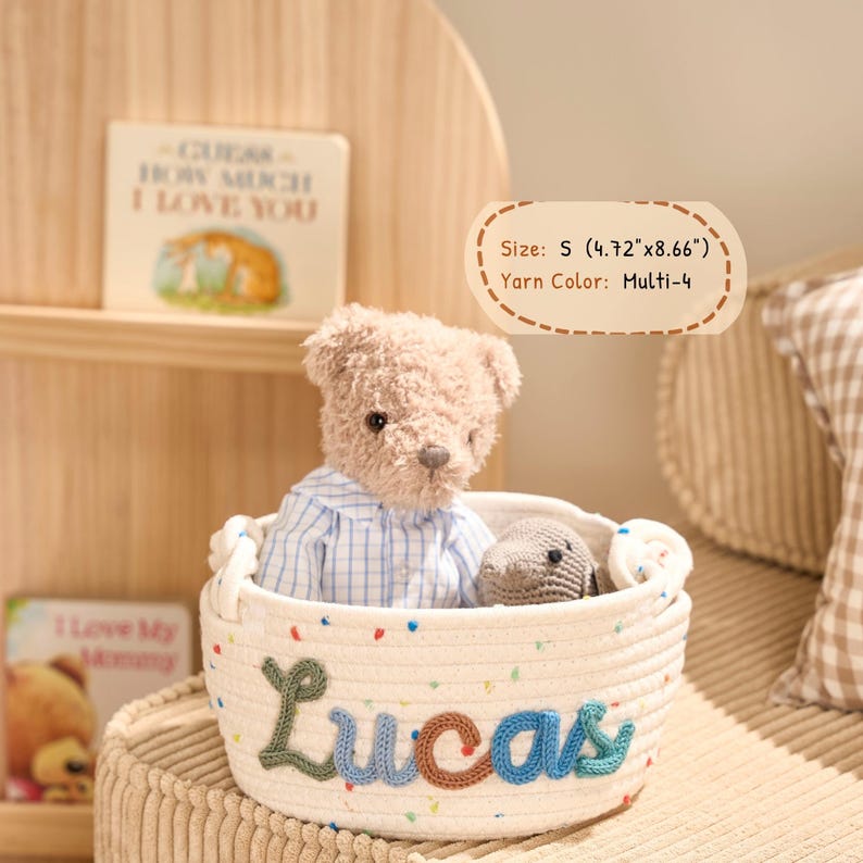 May include: A small, cream-colored rope basket with the name "Lucas" embroidered in green and blue yarn. The basket contains a teddy bear wearing a blue and white checkered shirt and a small gray crocheted animal. The basket measures 4.72" x 8.66".