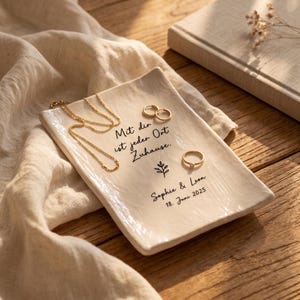 Könnte beinhalten: Eine rechteckige, cremeweiße Keramikschale mit glänzender, strukturierter Oberfläche. Sie ist mit dem schwarzen Schriftzug "Mit dir ist jeder Ort Zuhause.", einem kleinen Blatt-Symbol, "Sophie & Leon" sowie "18. Juni 2025" bedruckt. Eine goldene Kette und zwei Ringe liegen darauf.