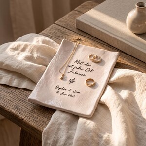 Könnte beinhalten: Eine rechteckige, cremeweiße Keramikschale mit glänzender Oberfläche. Die Schale ist mit dem schwarzen Text "Mit dir ist jeder Ort Zuhause.", "Sophie & Leon", "18. Juni 2025" und einem kleinen Blattsymbol bedruckt. Eine goldene Kette und drei goldene Ringe liegen auf der Schale.