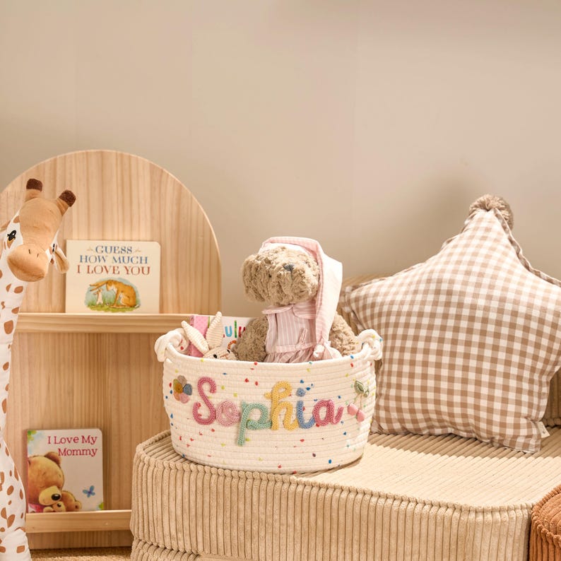 May include: A white rope basket with the name 'Sophia' embroidered in colorful letters. The basket contains a teddy bear wearing a pink dress and a bunny toy. A star-shaped pillow and a wooden shelf with books and a giraffe toy are in the background.