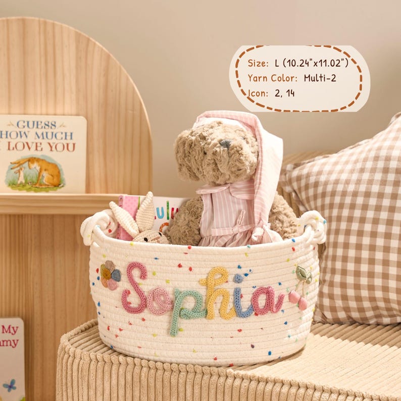 May include: A cream-colored woven basket with the name "Sophia" embroidered in colorful yarn. The basket contains a teddy bear wearing a pink nightcap and a small stuffed bunny. The basket is approximately 10.24" x 11.02".