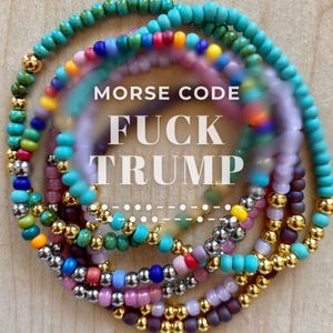 Peut inclure: Plusieurs bracelets de perles colorées, aux tons turquoise, violet, vert, jaune et or. Les bracelets sont disposés en cercle, avec le texte "MORSE CODE FUCK TRUMP" superposé à l'image.