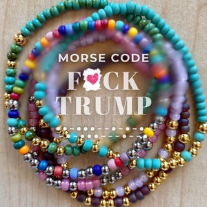 May include: A collection of circular beaded bracelets featuring a variety of colorful glass and metallic gold beads. The text MORSE CODE FOCK TRUMP is centered in white lettering over the bracelets, with a small pink heart icon above the word FOCK.