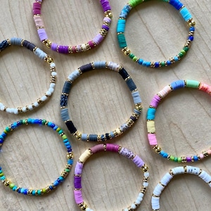 Puede incluir: Una colección de pulseras de cuentas coloridas. Cada pulsera presenta una variedad de colores, incluyendo azul, verde, rosa, morado y blanco, con cuentas doradas intercaladas. Las pulseras están dispuestas sobre una superficie de madera marrón claro.