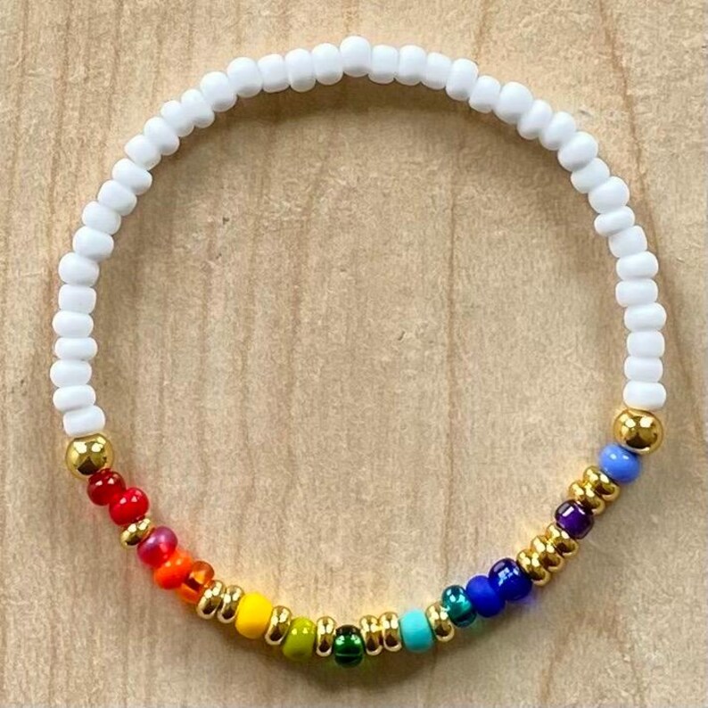 Pu&ograve; includere: Un braccialetto di perline colorate con un design arcobaleno. Il braccialetto presenta perline bianche e dettagli dorati, con una sequenza di perline rosse, arancioni, gialle, verdi, blu e viola. Il braccialetto &egrave; su una superficie di legno marrone chiaro.