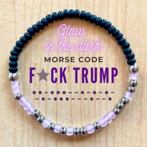 Peut inclure: Un bracelet de perles noires, argentées et lavande, avec le texte "Glow in the dark" et "Morse Code F*CK TRUMP". Le code Morse du texte est affiché en dessous.