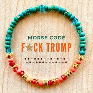 Op de afbeelding: Een kralenarmband met de tekst "MORSE CODE F*CK TRUMP". De armband heeft turquoise, koraal en gouden kralen. De Morsecode van de boodschap wordt eronder weergegeven.