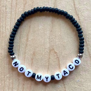 May include: Black beaded bracelet with white letter beads spelling "NOT MY TACO". The bracelet has silver accent beads. The bracelet is on a light brown wooden surface.