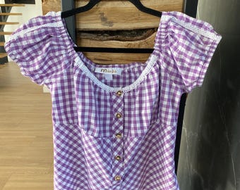 Puede incluir: Una blusa a cuadros morados y blancos con mangas cortas abullonadas y un escote cuadrado. La blusa tiene una fila de botones dorados en la parte delantera y ribete de encaje blanco. La marca "Marjo" es visible.