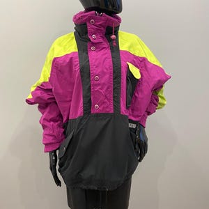 May include: A vintage anorak jacket in neon yellow, magenta, and black. The jacket has a high collar, snap closures, and a front pocket. The sleeves are wide and the jacket has a drawstring at the hem. A retro style, reminiscent of the 80s.