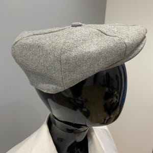 Könnte beinhalten: Graue Newsboy-Cap aus strukturiertem Stoff. Die Kappe wird auf einem schwarzen Schaufensterpuppenkopf präsentiert, kombiniert mit einem weißen Hemd mit Kragen. Die Kappe hat eine runde Krone und einen kleinen, steifen Schirm.