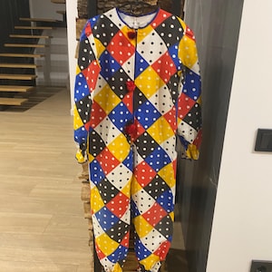 May include: A colorful, patterned jumpsuit with a harlequin design. The one-piece garment features a diamond pattern in red, yellow, blue, black, and white, with small white polka dots. It has long sleeves, a button-up front, and red pom-pom accents.