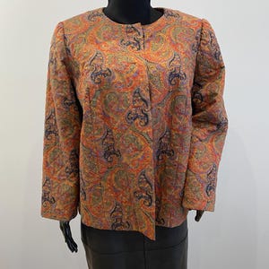Może przedstawiać: Kurtka w stylu vintage z nadrukiem paisley w odcieniach pomarańczu, czerwieni, zieleni i niebieskiego. Kurtka ma okrągły dekolt i długie rękawy. Kurtka jest prezentowana na manekinie.