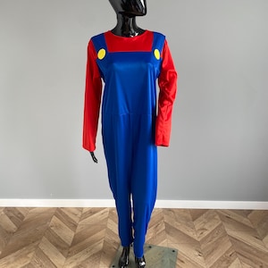 May include: A long-sleeved jumpsuit featuring a vibrant red shirt top and blue overalls. The blue section includes two large, circular yellow buttons positioned at the chest area. The garment is displayed on a black mannequin against a gray wall.