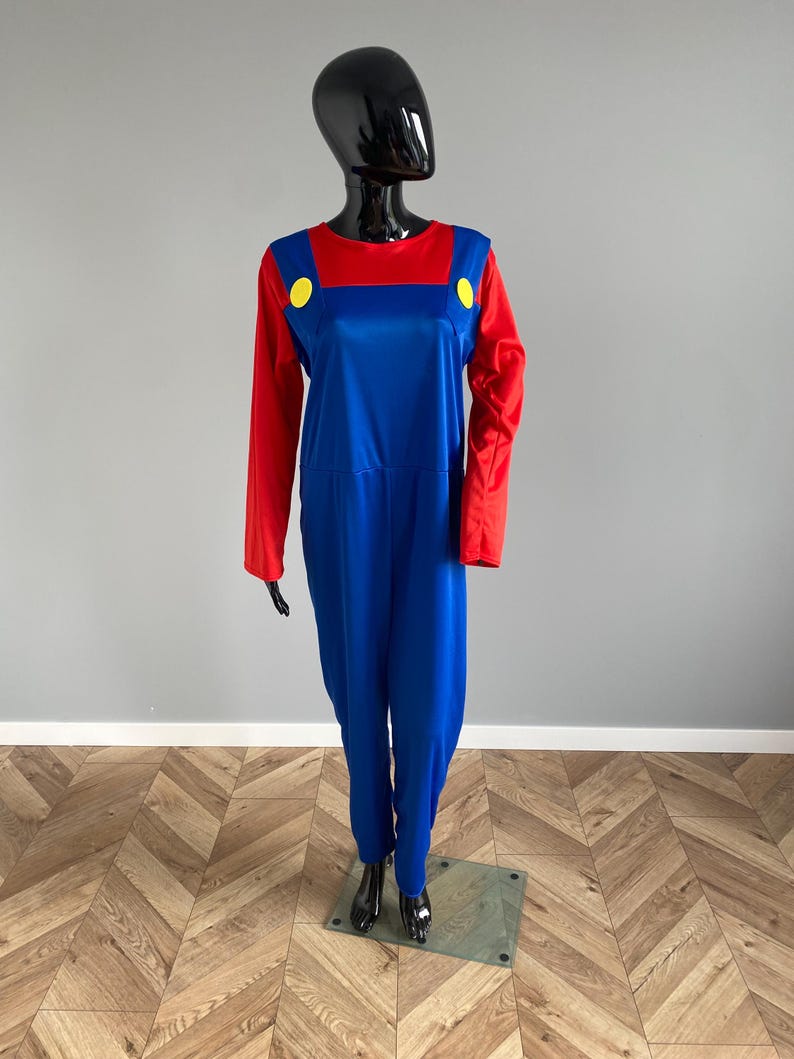 May include: A long-sleeved jumpsuit featuring a vibrant red shirt top and blue overalls. The blue section includes two large, circular yellow buttons positioned at the chest area. The garment is displayed on a black mannequin against a gray wall.
