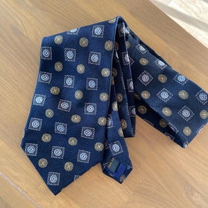 May include: Navy blue tie with a repeating pattern of squares and circles in shades of gold, white, and light blue. The tie is made of a smooth fabric and is laid out on a wooden surface.