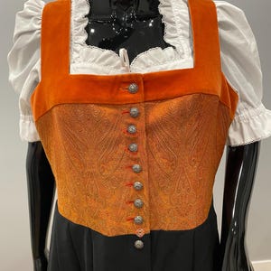 May include: An orange and gold paisley patterned bodice with silver buttons and red stitching. The bodice is paired with a white blouse with ruffled sleeves and a black skirt. The garment is displayed on a mannequin.