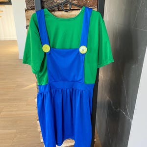 May include: A green t-shirt with blue overalls attached, featuring large yellow buttons. The bottom of the overalls is a pleated blue skirt. The costume is hanging on a black hanger.