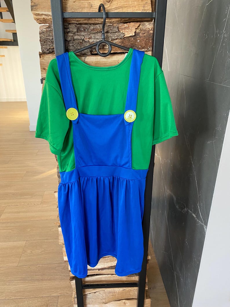 May include: A green t-shirt with blue overalls attached, featuring large yellow buttons. The bottom of the overalls is a pleated blue skirt. The costume is hanging on a black hanger.