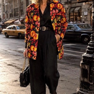 May include: A blazer featuring a vibrant, multi-colored fruit print on a black background, worn over a black collared shirt and black high-waisted trousers with a black belt, black pointed-toe heels, and a black handbag with a gold chain strap.
