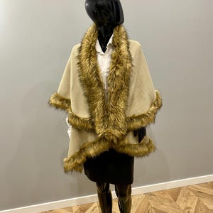 May include: A beige cape with faux fur trim and a matching fur collar is displayed on a mannequin. The cape is worn over a white collared shirt and a black skirt. The mannequin is wearing a black beret.