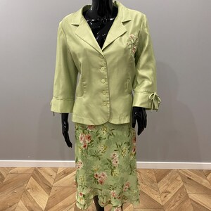 Op de afbeelding: Een lichtgroen tweedelig pak op een mannequin. Het pak bestaat uit een blazer met driekwart mouwen, een reverskraag en bloemenborduurwerk. De bijpassende rok heeft een bloemenpatroon met een gefranjerde zoom. Het pak wordt gepresenteerd op een zwarte mannequin.