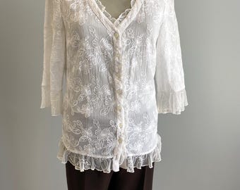 May include: A white, semi-sheer blouse with floral embroidery, a ruffled V-neck, and three-quarter sleeves. The blouse has a button-down front and a ruffled hem. Paired with dark brown trousers.