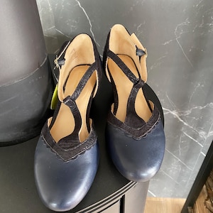 May include: A pair of navy blue leather T-strap shoes featuring a rounded toe, a scalloped edge detail, and a textured black fabric overlay on the upper. The shoes include a thin ankle strap with a small gold-toned buckle and a tan interior lining.