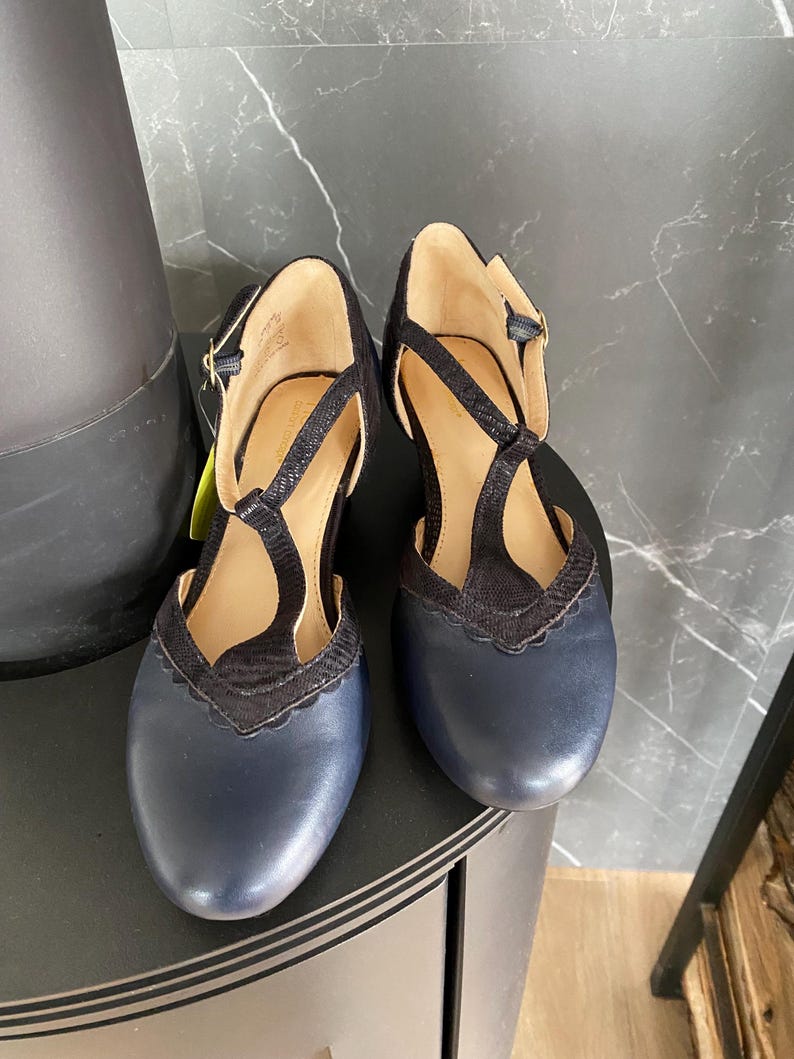 May include: A pair of navy blue leather T-strap shoes featuring a rounded toe, a scalloped edge detail, and a textured black fabric overlay on the upper. The shoes include a thin ankle strap with a small gold-toned buckle and a tan interior lining.