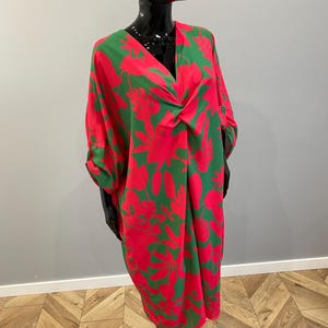 May include: A vibrant, knee-length dress with a bold floral pattern in red and green. The dress features a V-neckline and long sleeves that are rolled up. The fabric drapes loosely, creating a relaxed silhouette. The dress is displayed on a black mannequin.