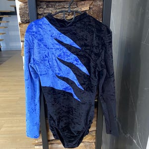 May include: A black and blue velvet leotard with long sleeves. The blue velvet is on the left sleeve and chest, with a jagged design. The leotard is hanging on a black metal rack.