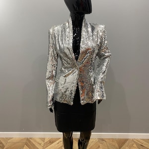 May include: A silver sequin blazer with a notched collar and long sleeves. The jacket is displayed on a black mannequin, paired with a black skirt. The blazer's shimmering sequins catch the light. The mannequin is standing on a wooden floor.