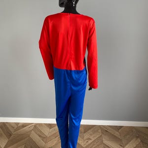 May include: A long-sleeved jumpsuit featuring a vibrant red bodice and bright blue trousers. The garment is constructed from a smooth, slightly reflective fabric and includes a vertical zipper closure centered along the upper back section.