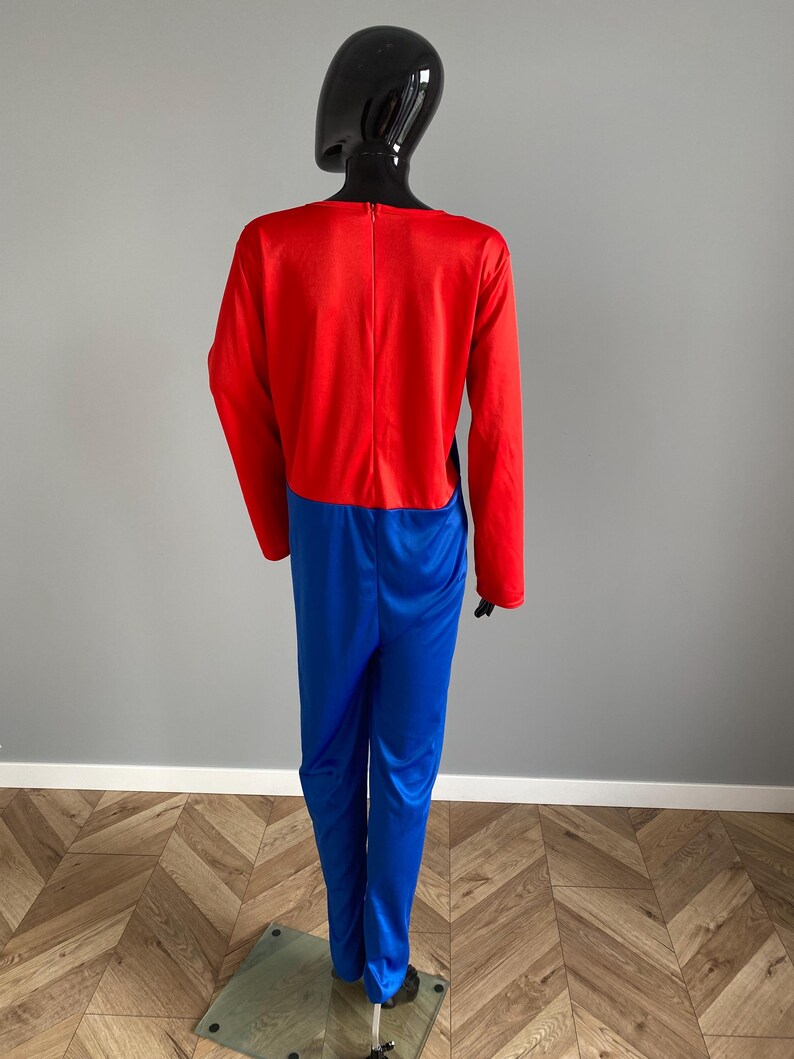 May include: A long-sleeved jumpsuit featuring a vibrant red bodice and bright blue trousers. The garment is constructed from a smooth, slightly reflective fabric and includes a vertical zipper closure centered along the upper back section.