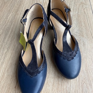 May include: A pair of navy blue T-strap shoes with a textured black overlay on the front and scalloped edges. The shoes feature a rounded toe, a buckle closure on the side strap, and a light-colored interior lining with a visible brand logo.