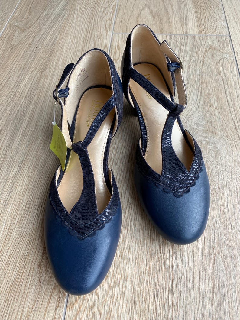 May include: A pair of navy blue T-strap shoes with a textured black overlay on the front and scalloped edges. The shoes feature a rounded toe, a buckle closure on the side strap, and a light-colored interior lining with a visible brand logo.
