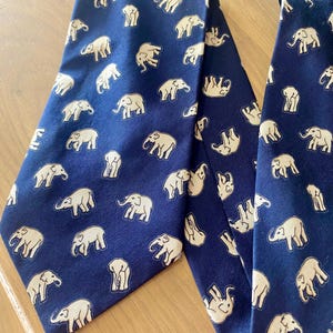 Op de afbeelding: Twee marineblauwe stropdassen met een herhaald patroon van cr&egrave;mekleurige olifanten. De stropdassen zijn gemaakt van een gladde stof en worden gepresenteerd op een lichtgekleurd houten oppervlak. Het olifantenontwerp is gedetailleerd en grillig.